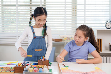 Asian girl and caucasian girl elementary classmates smiling while working on colorful crayon drawing at classroom desk representing creativity teamwork friendship childhood collaborative learning