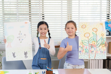 Asian girl and caucasian girl elementary classmates smiling holding artwork giving thumbs up gesture during classroom activity representing creativity pride friendship childhood artistic achievement
