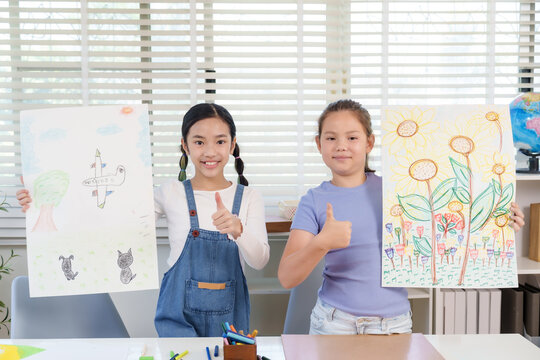 Asian girl and caucasian girl elementary classmates smiling holding artwork giving thumbs up gesture during classroom activity representing creativity pride friendship childhood artistic achievement