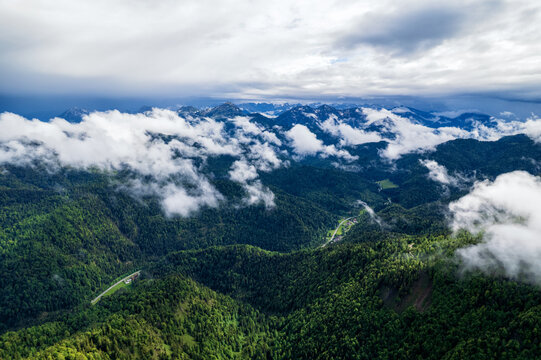 Aerial view of lush green forests meeting swirling clouds and distant peaks, a tapestry of nature's grandeur unfolds., Berchtesgaden, Bayern, Germany.