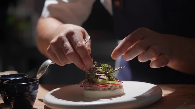 A chef is preparing a dish with a spoon and a knife. The dish is on a white plate and has a garnish of green leaves