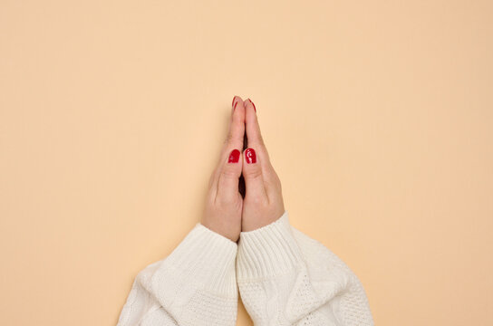 Female hands in a white knitted sweater, folded in a prayer pose or a gesture of gratitude. Beige background.
