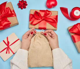 Female hands in a white sweater tightening a jute bag surrounded by Christmas gifts in craft paper...