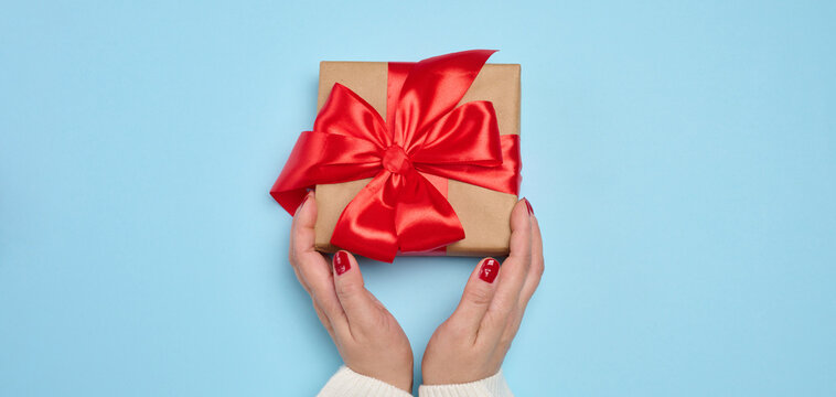 Two female hands holding a gift box wrapped in brown paper and a red ribbon on a blue background
