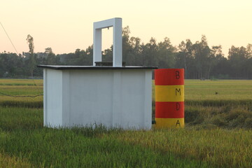 A small white structure with a red and yellow marker stands in a green field under a pale sky