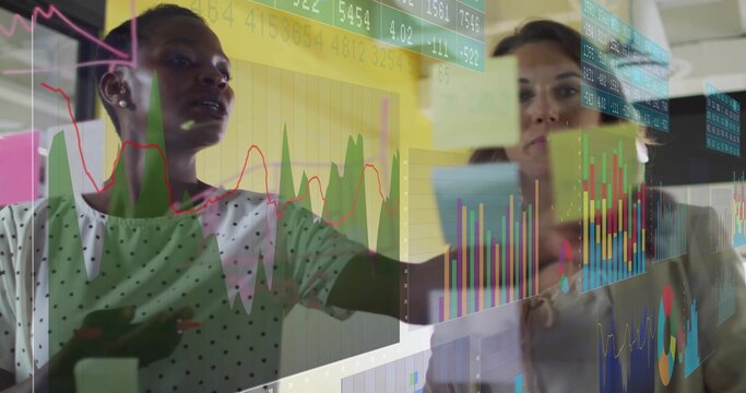 Pointing two women in polkadot and light tops analyzing glass planning wall with Postits and charts