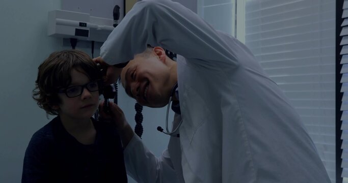 Leaning Asian doctor in lab coat examining child with otoscope on exam chair in clinic stethoscope