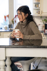 Contemplative woman holding a warm beverage while seated at a kitchen table in a cozy, well-lit home setting at midday