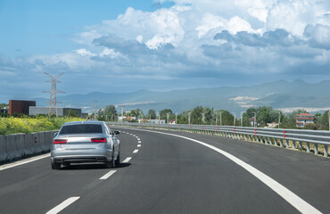 A car drives along the highway against the backdrop of countryside and mountains on a sunny day.