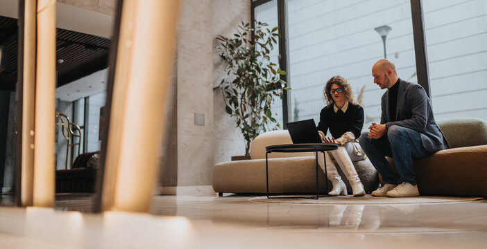 Two professionals sit on a contemporary lounge with a laptop, collaborating in a bright modern lobby, conveying teamwork, planning, and professional collaboration.