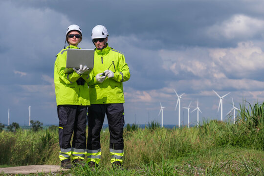 Two engineers checking data on a laptop near wind turbines under a cloudy sky in a renewable energy site