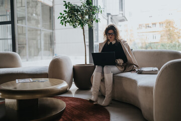 A stylish professional sits on a beige sofa in a modern lounge, typing on a laptop. Bright natural light and an urban backdrop suggest focus, remote work, and business collaboration.