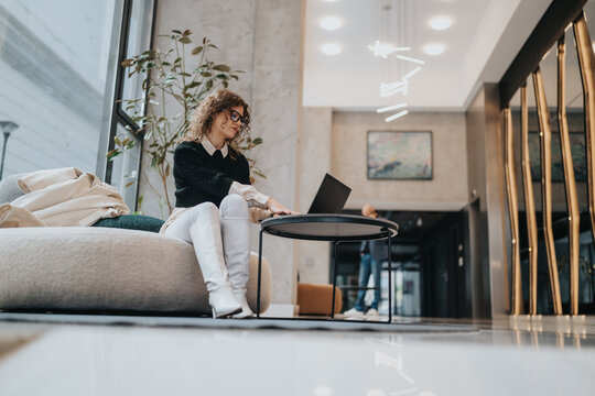 A stylish, professional woman sits on a comfortable sofa in a bright, modern lobby, working on her laptop at a round table. The calm, contemporary space suggests productivity and collaboration.