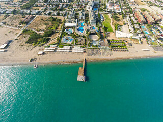 Aerial view of a wide sandy beach, turquoise sea, and a wooden pier extending out from a row of beachfront resort facilities and sun loungers