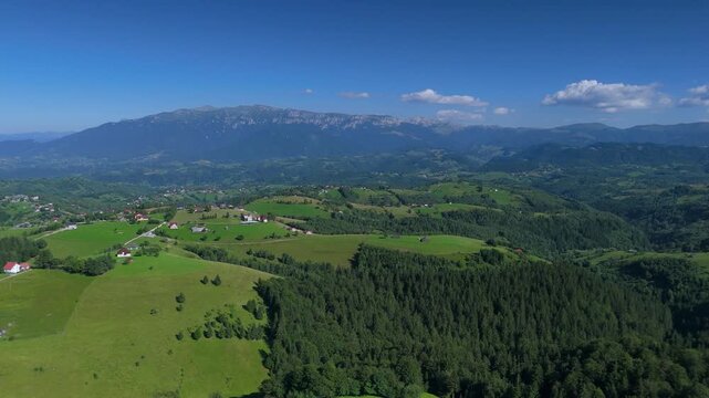 Panoramic view over the rolling green hills of the Bran Rucar corridor with Bucegi Mountains ridge under a clear summer sky, aerial footage in Transylvania, Romania, eco tourism, countryside travel