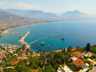 Wide aerial view showing the Alanya harbor pier, the Red Tower (Kızıl Kule) area, ships in the turquoise bay, and the extensive city stretching along the coast