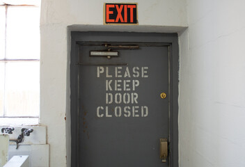 Gray Metal Exit Door with "Please Keep Door Closed" Stenciled Text and an Illuminated Exit Sign Above.
