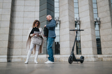 A man and woman in smart casual attire stand outside a contemporary office facade, reviewing a printed document while a black electric scooter sits nearby. © qunica.com