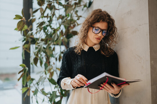 A thoughtful woman with curly hair and glasses reads a notebook in a modern office setting. Natural light, plants, and a calm, focused mood convey study and work themes.