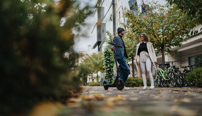 A man on a scooter chats with a woman in stylish outerwear on a tree-lined city street. Bicycles parked nearby and fallen leaves create a warm, urban autumn atmosphere.