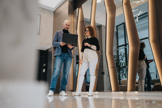 Two professionals stand in a sleek, reflective lobby with a laptop, engaged in a collaborative discussion. The scene conveys teamwork, business planning, and contemporary office style.