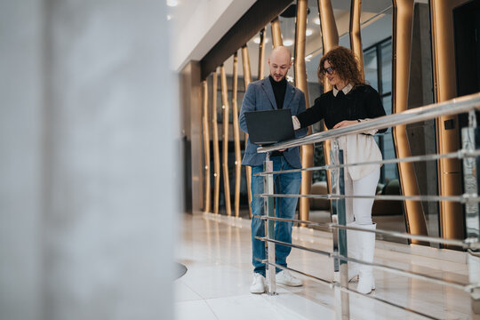 Two professionals stand by a railing in a bright, contemporary office, examining a laptop. They collaborate on ideas, planning and strategy, showcasing teamwork in a corporate environment.