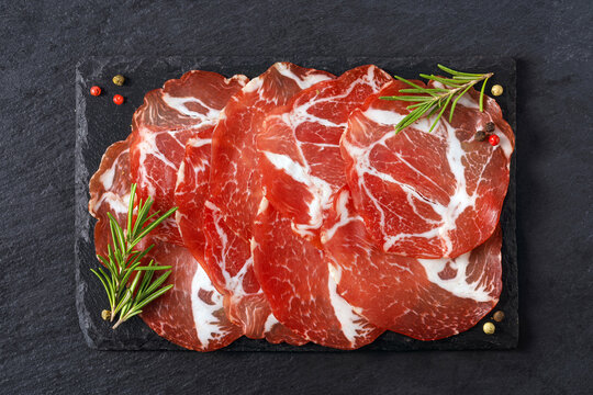 Sliced capicola coppa meat on dark slate board with rosemary