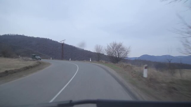 View from a moving vehicle showing a lake with a water tower in rainy weather and a winding mountain road with a sharp turn warning sign.

