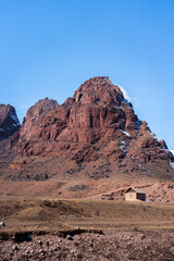 A tiny, isolated house sitting in a dry, barren landscape at the foot of a towering, reddish-brown rock mountain under a clear blue sky