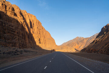 An empty, straight asphalt road cutting through a narrow valley, flanked by towering orange-red rock cliffs in a deep mountain gorge