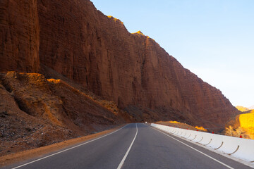 A straight asphalt road runs along the base of a colossal, textured red-brown cliff face in a mountain canyon under a bright sky