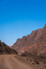 Fototapeta premium An unpaved road winding through a tight mountain pass or valley framed by massive, rugged red rock cliffs under a bright blue sky
