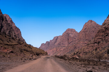 An unpaved road winding through a tight mountain pass or valley framed by massive, rugged red rock...