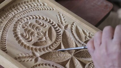 Close-up of an artisan's hand using a chisel to carve intricate floral ornaments into a wooden board, showcasing traditional woodworking skills and precise attention to beautiful artistic details.

