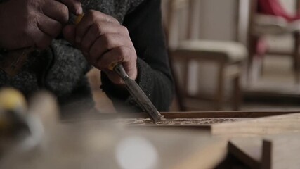 Close-up of an artisan's hand using a chisel to carve intricate floral ornaments into a wooden board, showcasing traditional woodworking skills and precise attention to beautiful artistic details.

