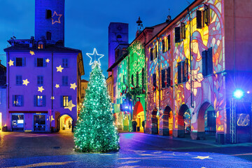 A Christmas tree stands on a town square surrounded by historic buildings, illuminated in the evening with festive light projections, in Alba, Italy.