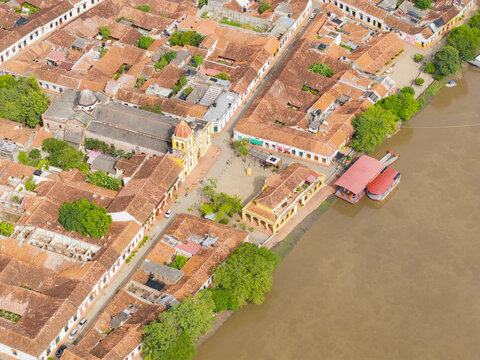 Aerial view of terracotta rooftops meet the Magdalena muddy river's edge, framed by the church's pale facade and the vibrant green of the trees, Mompox, Santander, Colombia.