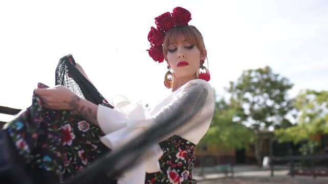 Graceful woman with red flowers in her hair dancing flamenco. Passionate artist in a traditional spanish costume performing outdoors with a fan