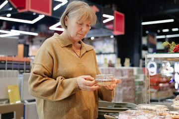A woman in a cozy sweater looks thoughtfully at a dessert container while shopping in a stylish bakery filled with various treats