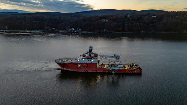 Aerial view of a vibrant red offshore supply vessel cuts through the calm, dark waters of the Hudson River, framed by the forested banks, Poughkeepsie, New York, United States.