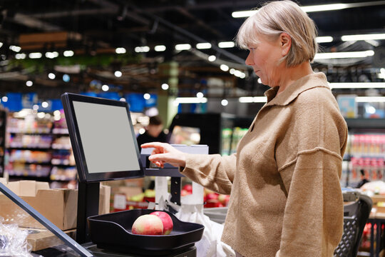 A woman is at a grocery store self-service kiosk weighing apples. She focuses on the screen, ensuring her purchase is calculated accurately