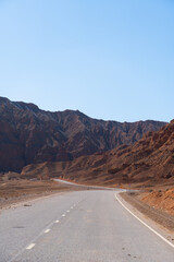 A long, empty asphalt road winding through a wide, arid valley surrounded by towering, red-brown mountains under a vast clear sky