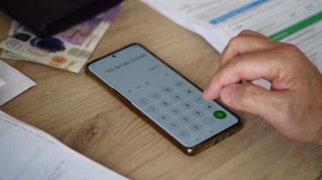 A person adding up the cost of domestic expenses by using a phone calculator on a desk to calculate household bills. A cost of living concept.