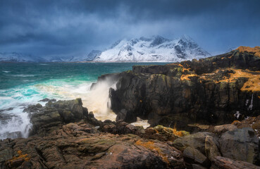 Wave impact on a rocky coastline with turquoise sea and snowy mountains in Lofoten Islands, Norway. Dynamic coastal landscape with cliffs, winter colors and rough water. Dramatic scenery with rocks