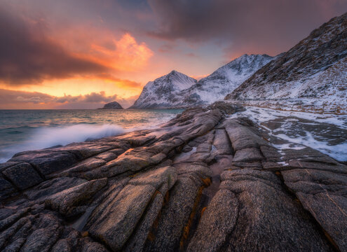 Dramatic winter seascape on the Lofoten Islands in Norway with waves crashing over coastal rocks and snow-covered mountains at sunset. Arctic landscape with sea, stones, blurred water, colorful sky