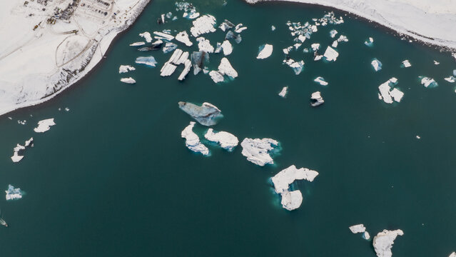 Aerial view of glacial lagoon speckled with luminous icebergs drifting in dark water under a dusting of snow, Jokulsarlon, Sveitarfelagid Hornafjordur, Iceland.