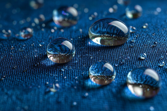 Gotas de agua sobre tela azul con textura visible, mostrando alta tensi&oacute;n superficial y efecto hidrof&oacute;bico que impide la absorci&oacute;n, ideal para tejidos t&eacute;cnicos y prendas resistentes al agua.