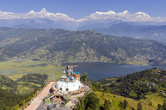 Aerial view of the majestic Shiva statue perched atop a hill overlooking the serene Phewa Lake, with the snow-capped Annapurna range piercing the skyline, Pumdikot, Gandaki Province, Nepal.