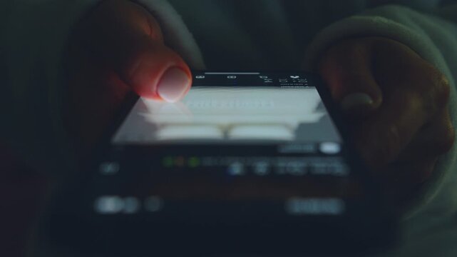Close-up of a person's hands using a phone, holding a smartphone and browsing social media. The screen glows softly, reflecting off the user's fingers. 