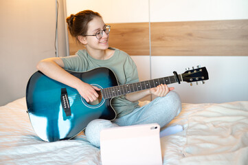 A teenage girl plays the guitar, showing it to someone online while smiling, illustrating joy of music, digital interaction, and engaging leisure activities at home